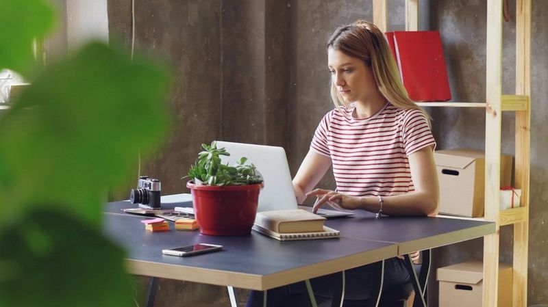 Kırmızı ve beyaz çizgili tişörtü olan bir kadın, masada oturuyor ve dizüstü bilgisayarda çalışıyor.