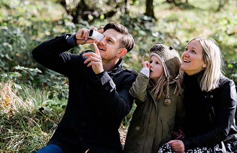 A young girl in a woolly hat sits with her parents in the woods, all three looking at something out of shot as her father uses a Canon PowerShot ZOOM.