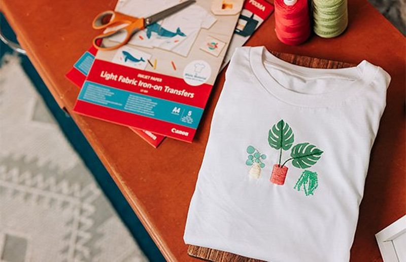 A white T-shirt with a printed embroidery pattern of houseplants, beside a pack of Canon iron-on transfers.