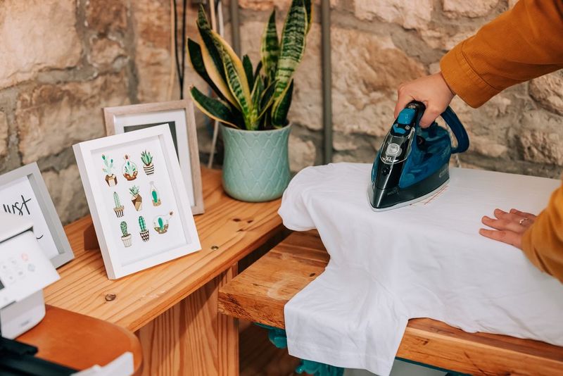A woman moving an iron across a T-shirt and a transfer design placed facedown.