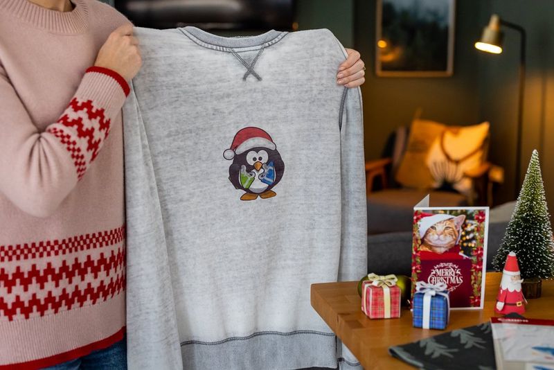 Жена држи сив џемпер украсен со слика од празничен пингвин што била пренесена врз џемперот со пеглање.