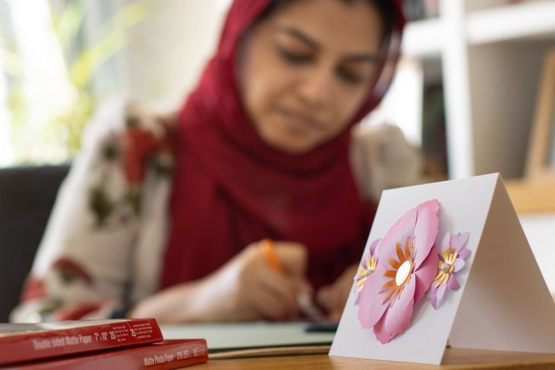Завршена картичка со тродимензионален цвет е поставена на маса покрај две кутии со хартија за печатење од Canon. Во заднината се гледа жена којашто изработува картички.