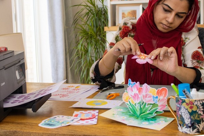 Жена внимателно држи цвет од хартија над маса прекриена со шарени рачно изработени картички, прибор за изработување на картички и печатач.