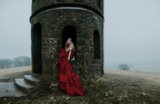 A woman with long blonde hair wearing a red gown sits in a castle window on a misty day. Taken by Rosie Hardy. 