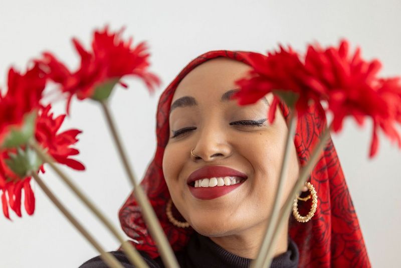 A woman smiles for the camera with her eyes closed. The bunch of red flowers she is holding partially obscures her face.