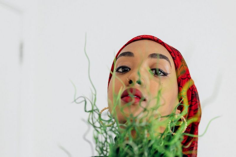 A close-up portrait of a woman in a red headscarf holding a plant up against her face.