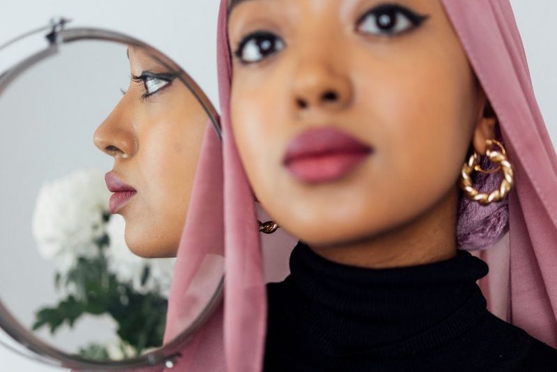 A self-portrait of photographer and poet Amaal Said in a pink headscarf. Her face is reflected in a small, round mirror she is holding.