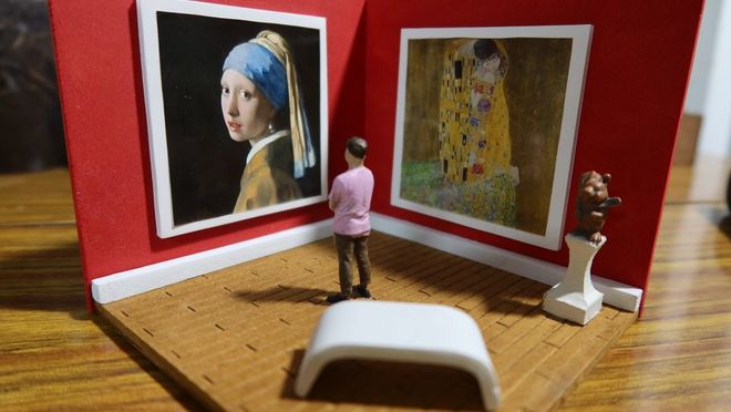 A mini papercraft art gallery with two paintings hung on red walls, and a mini figure standing in the foreground, looking at the art.