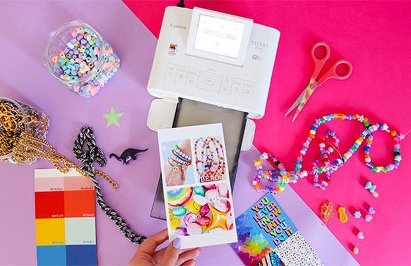 A hand taking a printed sheet of vibrant images from a Canon SELPHY CP1300 printer surrounded by beads, necklaces and a colour chart.