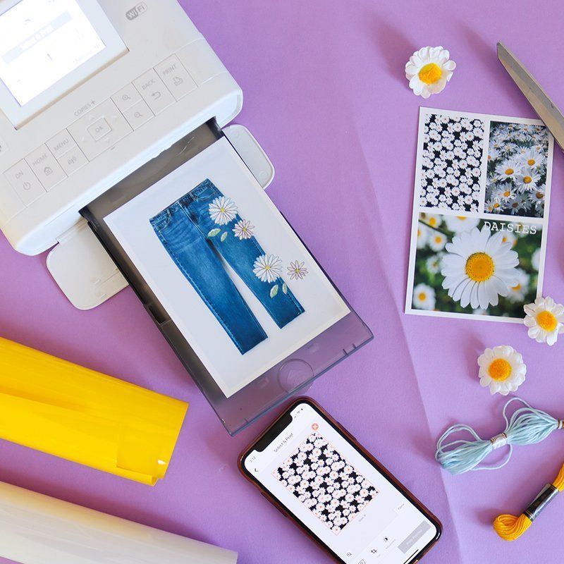 White SELPHY CP1300 printer with a picture of jeans and daisies printed out. Next to it there's a picture with 3 images on it and a smartphone with a black and white design image.