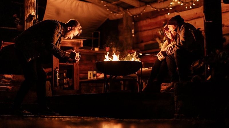 Un fotograf filmează doi oameni în haine de iarnă, cuibărindu-se unul lângă altul în timp ce coc bezele la un foc deschis. Scena este luminată doar de foc şi de un şirag de luminiţe în fundal.