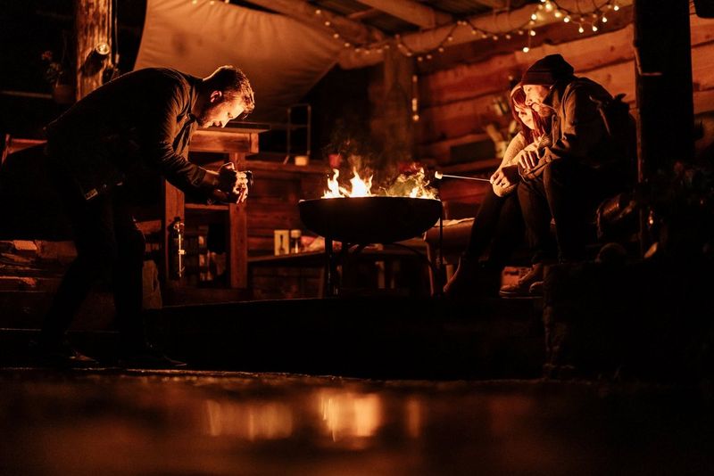 A photographer films two people in winter clothing, huddled together as they toast marshmallows on an open fire. The scene is lit solely by the fire and a string of fairy lights in the background. 