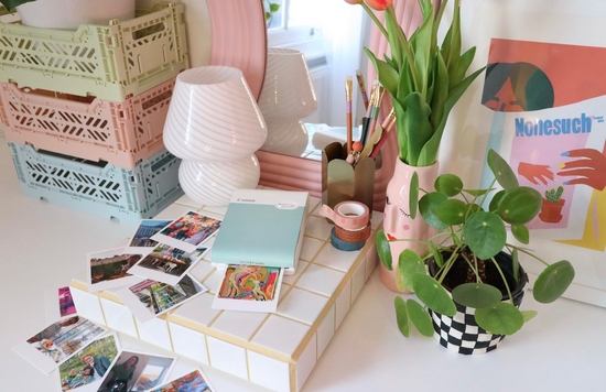 A top-down image of a Canon SELPHY Square QX10 printer on a desk surrounded by mini prints, stationery, a small lamp, a vase of flowers and a plant. 