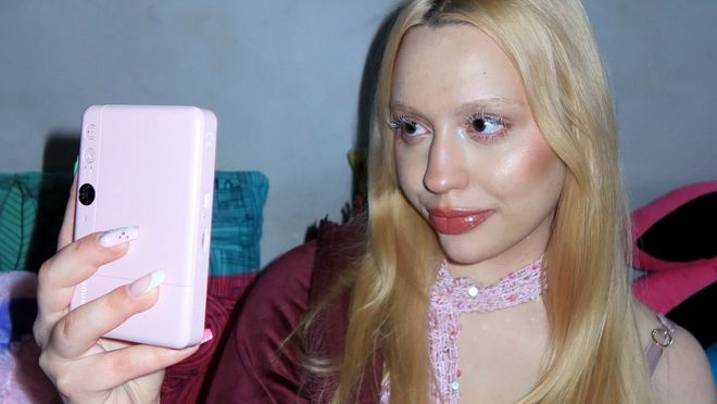 Style influencer Molly Wójcik, in een bordeauxrood jack en sjaal met lovertjes, houdt een Canon Zoemini S2 in roségoud omhoog om een selfie te maken.