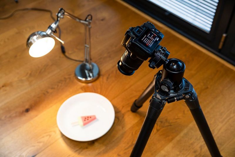 A Canon EOS 250D set up on a tripod facing an ice cream shaped like a slice of watermelon on a plate, with a desk lamp to one side.