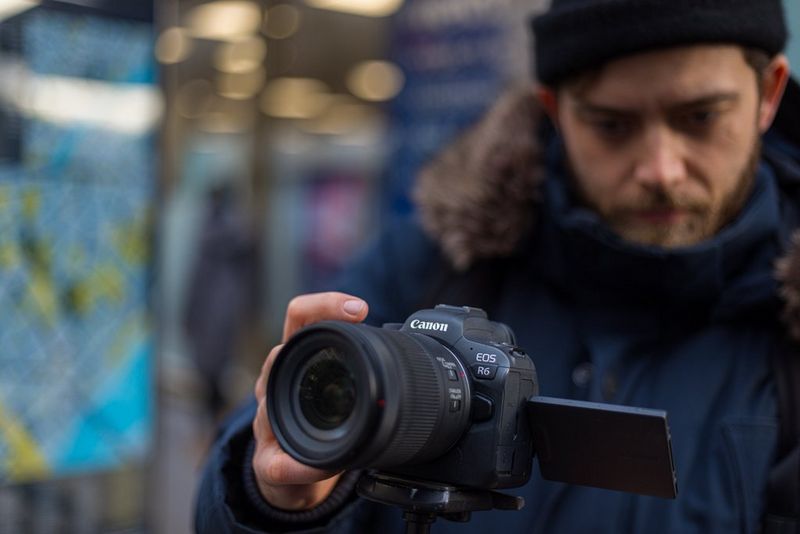 Мужчина в пальто и шерстяной шапке настраивает параметры Canon EOS R6, установленной на штатив.