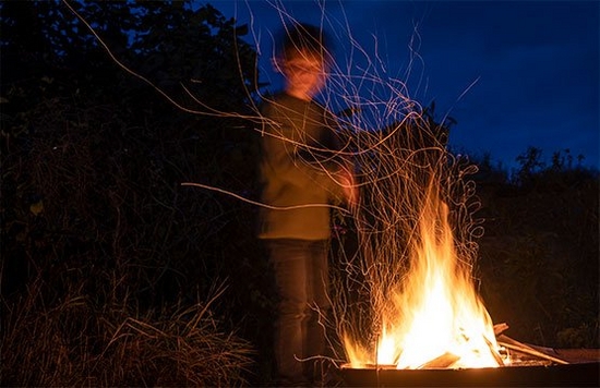 A long exposure of a child standing beside a bonfire, with floating embers creating long light trails.