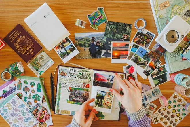 يدان تلصقان صورًا في دفتر يوميات رحلات سفر مفتوح محاط بصور وشريط وأغراض مؤقتة.