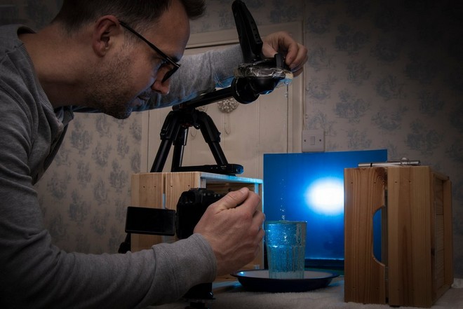 Egy fotós a fényképezőgépét kék lapnak támasztott, vízzel teli pohár fölé tartja, így kapja lencsevégre az aláhulló vízcseppet.