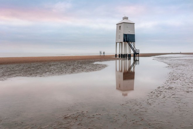 Vízimentő tornyot ábrázoló tengerparti látkép, amely a homokon lévő vízben tükröződik vissza.