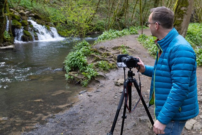 A fotós vízesésbe torkolló folyó előtt áll, fényképezőgépét állványra helyezve. 