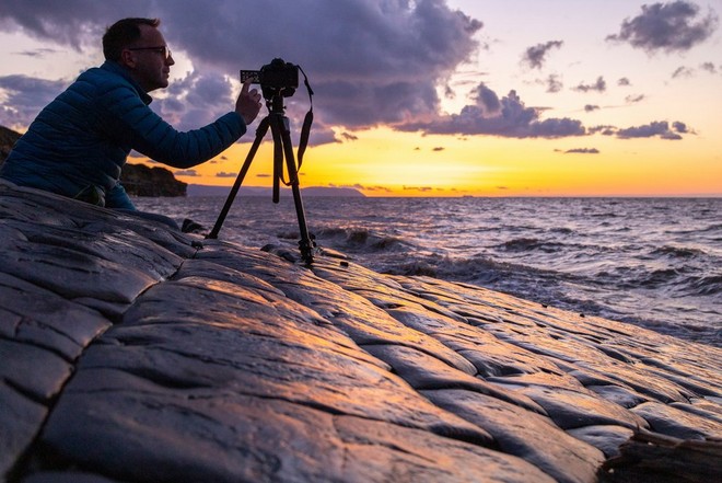 Egy kavicsos tengerparton felállított állványra helyezett fényképezőgép mellett guggoló férfi.