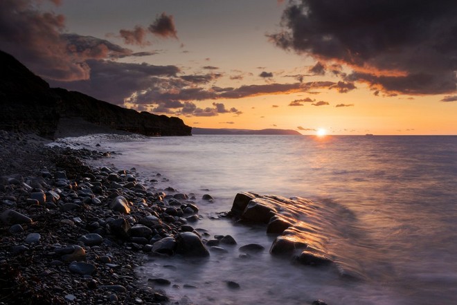Hosszú expozícióval készült kép a naplementében fürdő tengerről, amely elhomályosodik a partvonal mentén álló szikláknál.