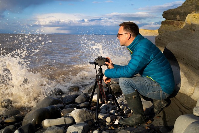 Egy fotós a tajtékzó hullámok melletti sziklákon álló állvány és fényképezőgép mellett guggol.