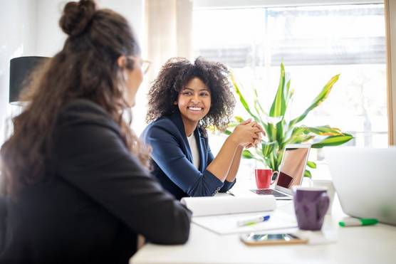 Woman smiles at a colleague while both sat at a white desk with a bright green plant.