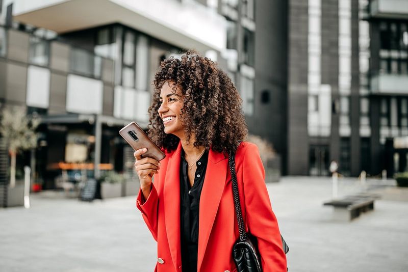 A woman wearing red, standing outside a building, holding her phone and smiling.