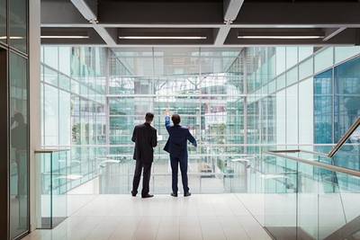 Two suited men with their backs to the camera gaze out through a large glass office building, with one pointing upwards.
