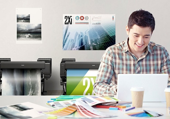 Man wearing a check shirt sitting at a white laptop, surrounded by two digital printers and colour charts spread out on the desk.