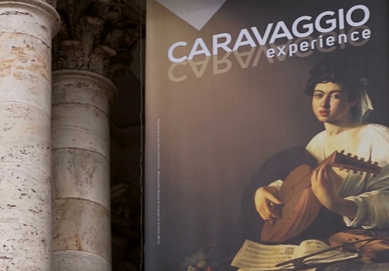 Two light-coloured stone pillars with a banner advertising the CARAVAGGIO experience, with an image of one of the artist’s paintings – a woman dressed in white playing a lute.