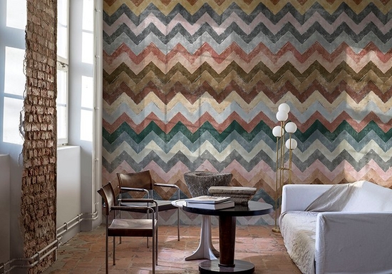 Image of a room with one wall exposed brick, and one covered with digitally printed wallpaper in multi-coloured chevrons, a round table, two chairs and a white sofa.