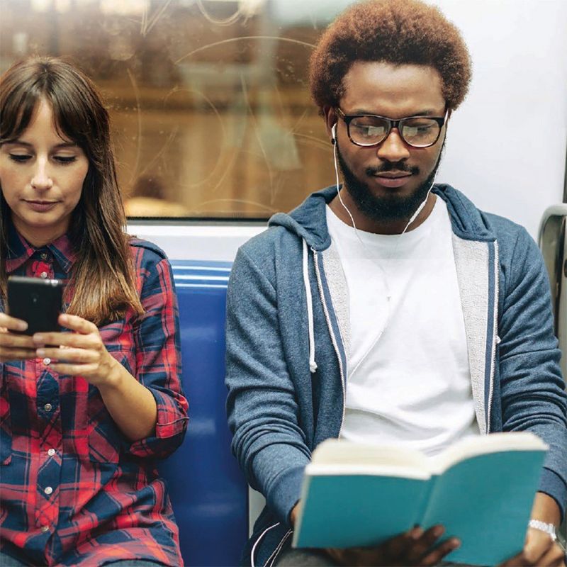 A woman in a check shirt looks at her smartphone and a man wearing a hoodie and white earphones reads a book on a train