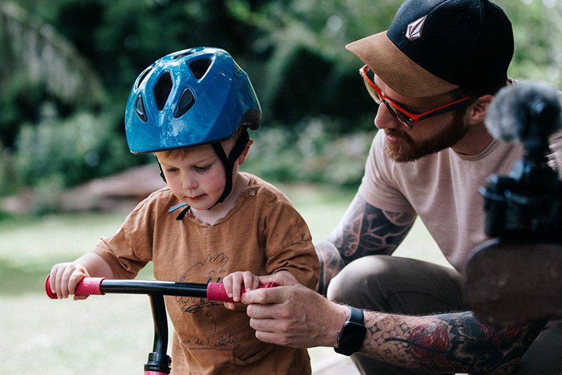 Stef Michalak helping his son hold onto the handles of his balance bike. Stef's EOS M50 is resting on the arm of a bench. 
