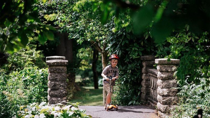Stef Michalak's eldest son, Grayson, riding a scooter over a bridge in a park. 