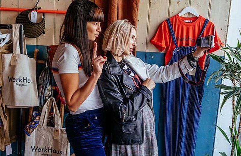 Two women pose in front of a camera in a clothes shop.
