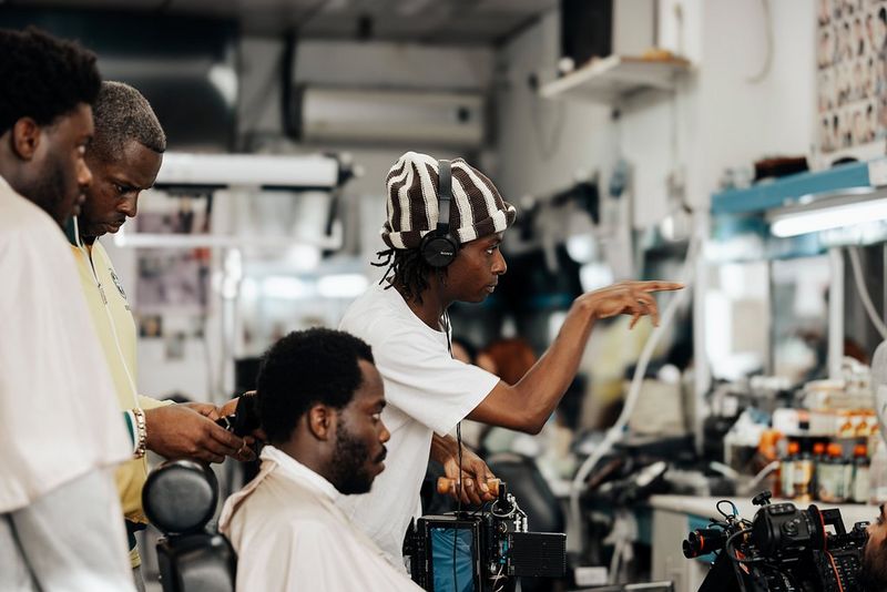 Filmmaker Samona Olanipekun wears headphones and points while an actor sits in a chair next to him and two other men stand behind the chair.