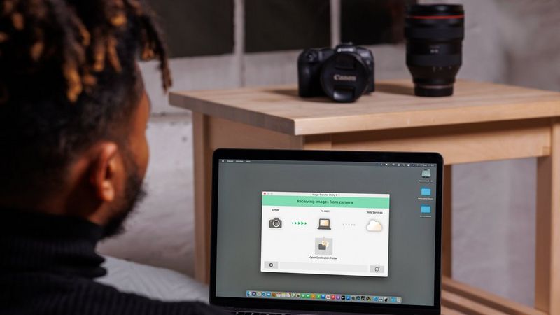 A black man looking at a screen of a laptop showing pictures being transferred from camera to computer using Image Transfer Utility software. Camera and lens visible in the background on a table. 