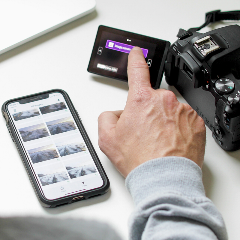 Smartphone with a picture on it, being held by a woman sitting at a table with camera in the background.