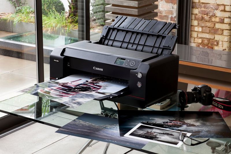 Une imprimante Canon imagePROGRAF PRO-1000 est posée sur une surface en verre, avec une photo récemment imprimée sur son plateau et d'autres impressions sur le côté.