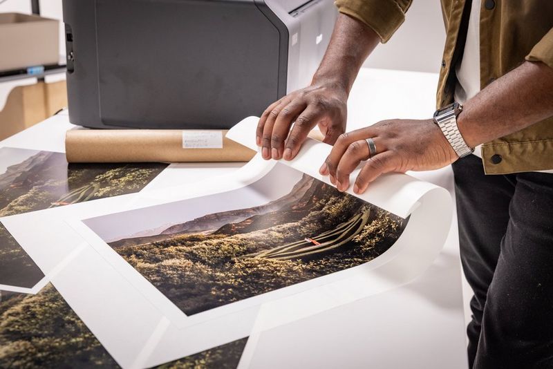 La main d'un homme commence à enrouler l'extrémité d'un tirage de paysage posé au sommet d'une pile d'autres tirages sur une table blanche, avec une imprimante Canon en arrière-plan.