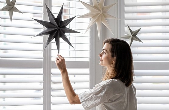 A woman touches a paper star hanging from her ceiling, in front of shuttered windows.