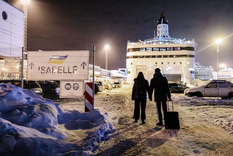 The silhouette of a couple holding hands and dragging a suitcase, as they walk through the snow towards a ferry under bright floodlights. To their left is a sign with arrows reading “ISABELLE”