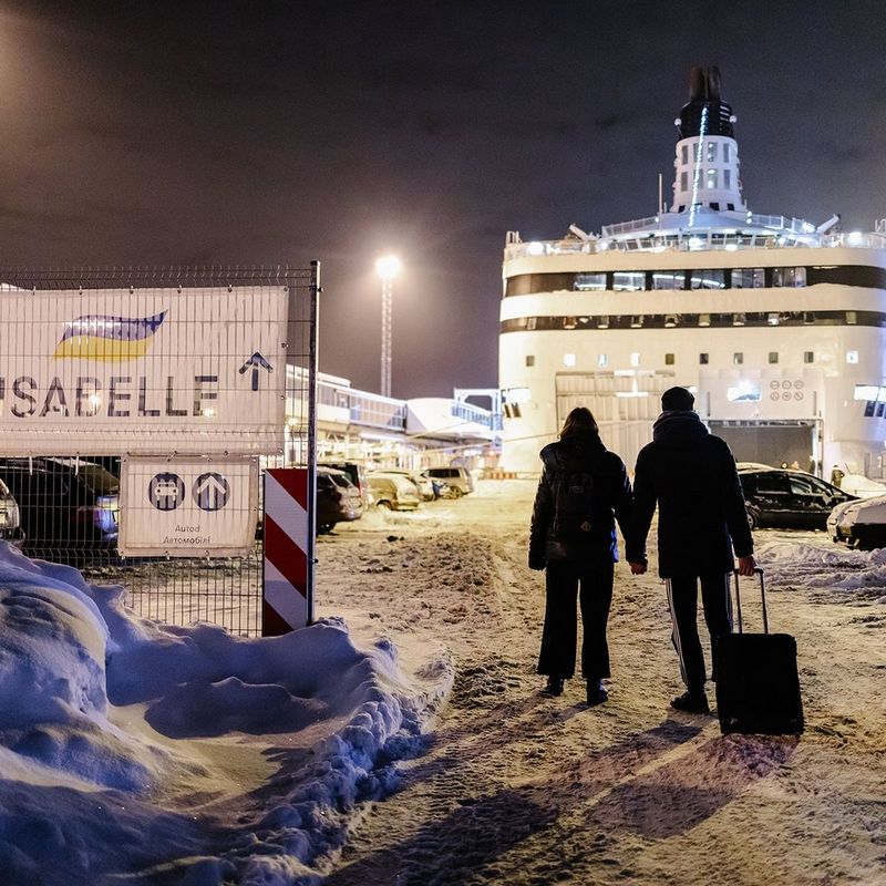 The silhouette of a couple holding hands and dragging a suitcase, as they walk through the snow towards a ferry under bright floodlights. To their left is a sign with arrows reading “ISABELLE”