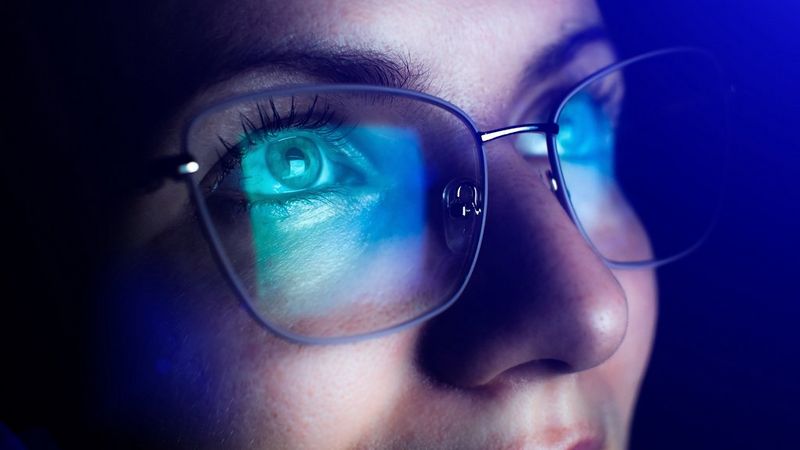 A womans bespectacled eyes and nose, bathed in blue light from a computer screen.