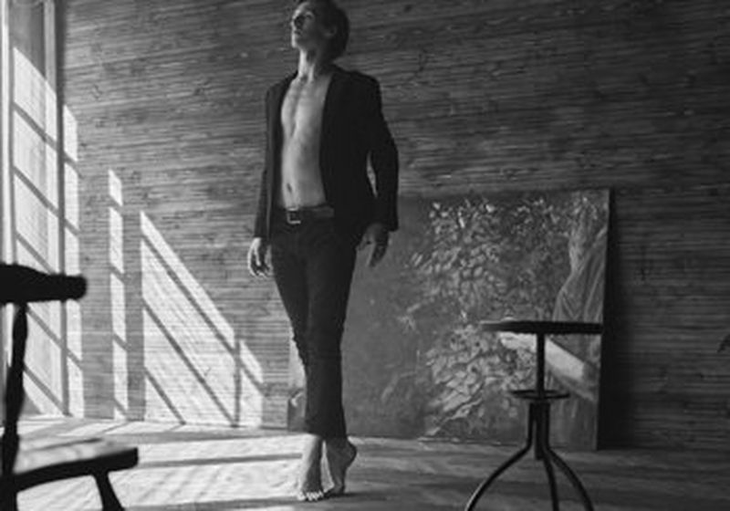 A black and white image of a barefoot ballet dancer en pointe, looking to the ceiling, in a sparsely decorated wood-walled room. Sunlight falls through the window. He wears jeans and an open blazer but is bare chested. Copyright Jaroslav Monchak