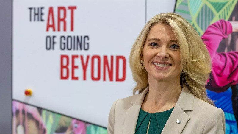 Jennifer Kolloczek is smiling and standing in front of a sign which reads ‘The Art of Going Beyond’