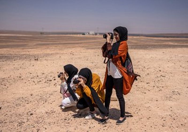 Three young women in headscarves stand in the desert. Two are crouching, one is stood. They are all holding cameras up to their faces and taking photographs.